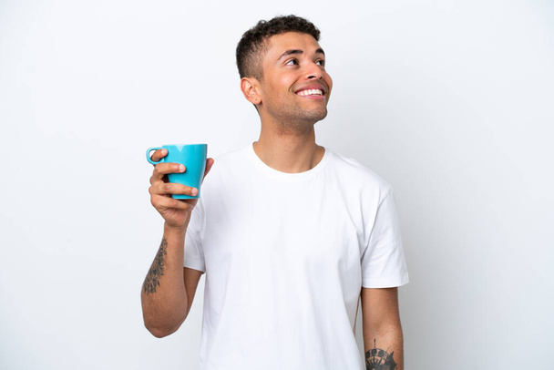 Young Brazilian man holding cup of coffee isolated on white background looking up while smiling - Фото, изображение