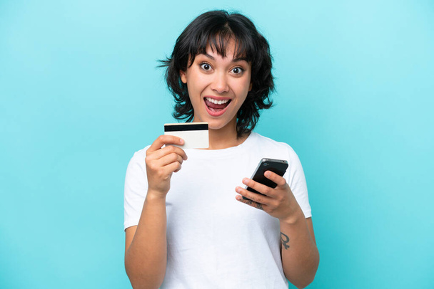 Giovane donna argentina isolato su sfondo blu acquisto con il cellulare e in possesso di una carta di credito con espressione sorpresa - Foto, immagini