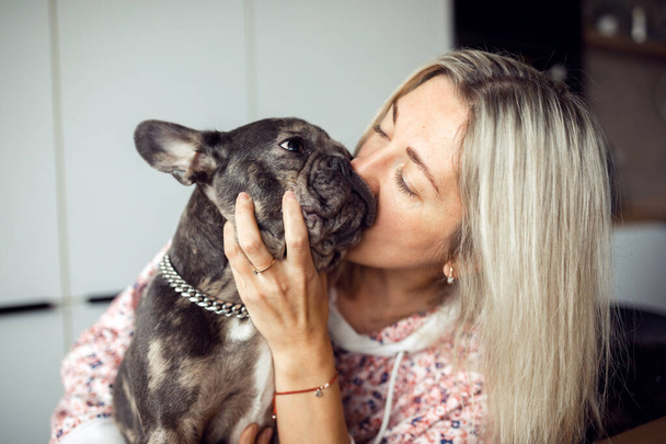Blonde woman he adores and kisses his French bulldog in cute muzzle. Maintenance and care of dogs. Healthy pets. Happy moments. Positive emotions. Pets. Friendship and mutual understanding.  - Fotó, kép