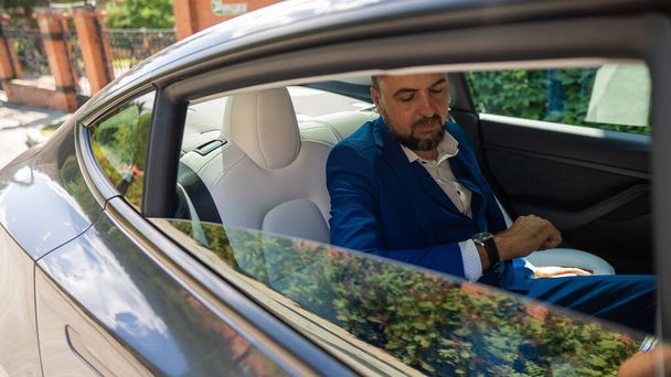 A caucasian man in a blue suit looks at his watch while sitting in the back seat of a car. Business class passenger - Фото, изображение