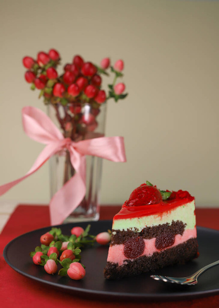 un pedazo de delicioso dulce hermoso pastel rayado brillante con fresas en un plato redondo blanco con una cuchara de postre calado y corazones junto a un jarrón con flores y un lazo - Foto, Imagen