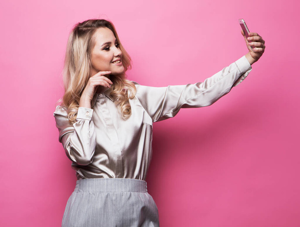 Blond pretty woman taking photo makes self portrait on smartphone over pink background - Foto, Imagen