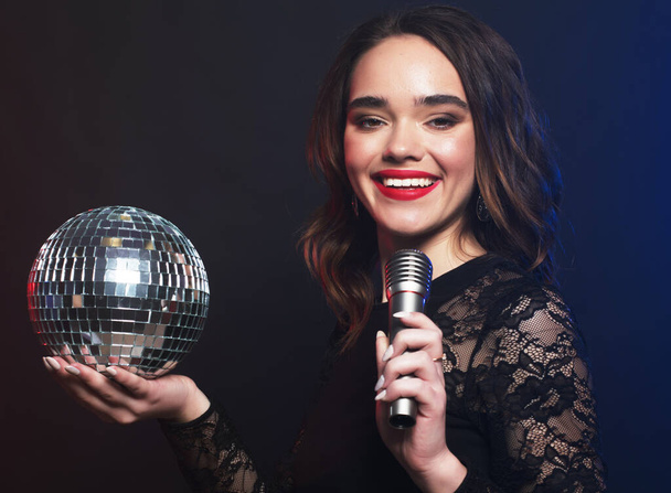 Lifestyle, party and people concept: young woman wearing black dress, holding disco ball and singing into microphone over dark background - Фото, изображение