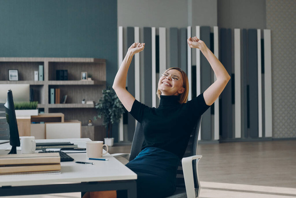 Excited young businesswoman keeping arms outstretched while sitting at the office desk - Фото, зображення