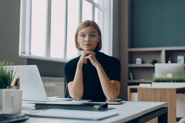 Confident businesswoman looking at camera while sitting at her working place in office - Фото, изображение