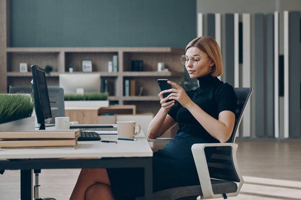 Αυστηρή νεαρή επιχειρηματίας που χρησιμοποιεί έξυπνο τηλέφωνο ενώ κάθεται στο χώρο εργασίας της στο γραφείο - Φωτογραφία, εικόνα