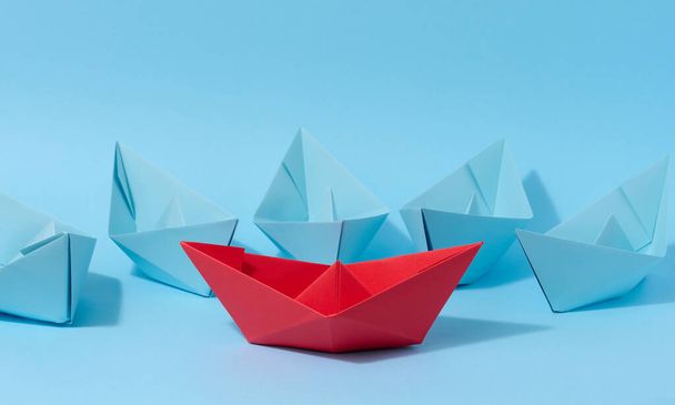 A red paper boat stands in front of a group of blue paper boats, a confrontation - Fotoğraf, Görsel