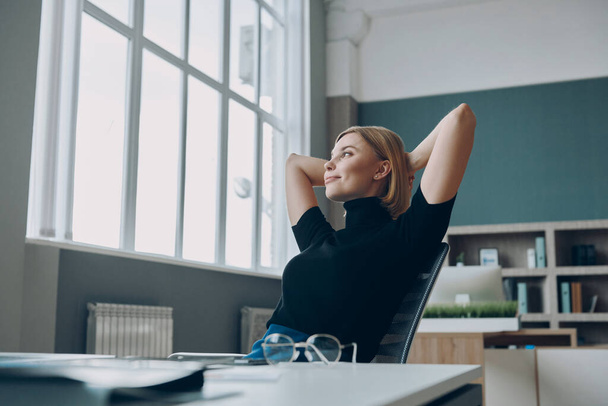 Jovem empresária de mãos dadas atrás da cabeça enquanto descansa em seu local de trabalho no escritório - Foto, Imagem
