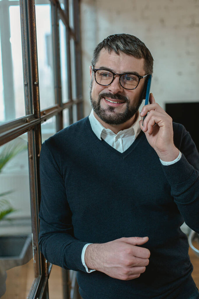 Красивый взрослый мужчина разговаривает по мобильному телефону и улыбается стоя в офисе - Фото, изображение