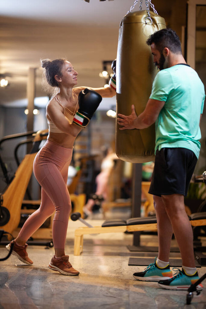 Fit girl with boxing gloves hitting a punching bag. Male fitness instructor with a female client practicing kickbox in a fitness studio. - Valokuva, kuva