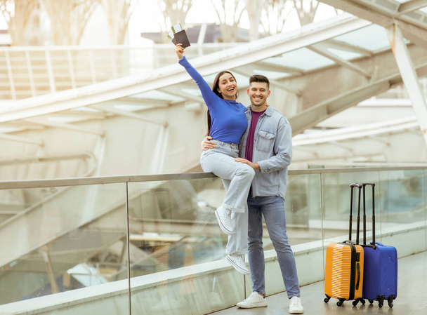 Podróże wakacyjne. Radosni turyści para pozowanie posiadania paszportu i kart pokładowych stojących z walizkami uśmiechnięty do kamery w nowoczesnym terminalu lotniska w pomieszczeniach. Strzał o pełnej długości - Zdjęcie, obraz