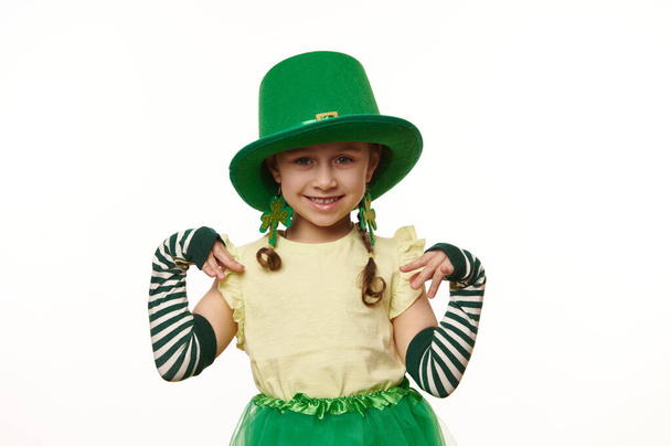 Απομονωμένο πορτρέτο σε λευκό φόντο ενός αξιολάτρευτου χαμογελαστού μικρού κοριτσιού, ντυμένο σαν ξωτικό με πράσινο καπέλο καρναβαλιού και ριγέ γάντια, έτοιμο για ένα πάρτι της Ημέρας του Αγίου Πατρικίου - Φωτογραφία, εικόνα