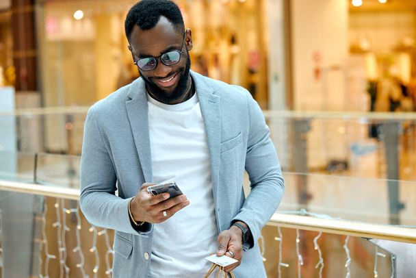 excited guy in eyeglasses looking at the screen of his smartphone,handsome man in fashion blue suit and black trousers searching Sales And Special Offers Standing indoors at Mall Store. - Zdjęcie, obraz