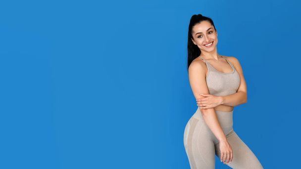 Pretty athletic young brunette woman in tight beige sportswear looking at free space for advertisement and smiling on blue studio background, web-banner for women workout - Photo, Image
