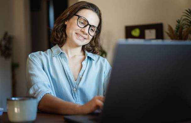 Happy creative young woman in eyeglasses working on laptop at home. Remote work, small business concept - Фото, изображение
