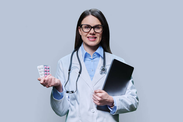 Young woman doctor recommending different pills in blister pack, female smiling holding laptop looking at camera on grey background. Medicine, healthcare, treatment, pharmacy, pharmacology concept - Photo, image