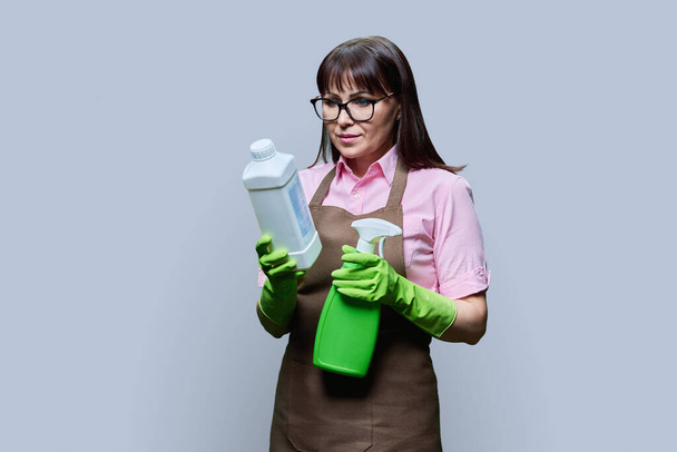 Middle aged woman in gloves apron with organic detergents, in hands looking at bottle on grey color background. Home cleaning service, housekeeping cleanliness, natural eco organic detergents, ecology - Photo, Image