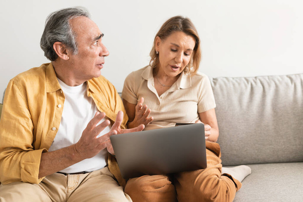 Couple âgé mécontent argumenter, en regardant leur compte bancaire en ligne sur ordinateur portable, femme en utilisant l'ordinateur et les achats en ligne à la maison, espace libre - Photo, image