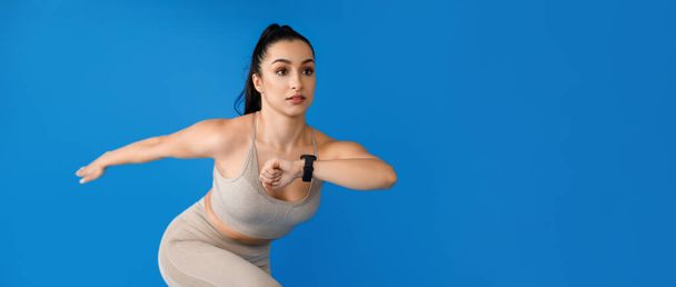 Мотивированная молодая спортсменка в обтягивающей бежевой спортивной одежде, бегущая по голубой stuido фона, женщины имеют тренировку, используя умные часы для фитнес-отслеживания, панорама с копировальным пространством - Фото, изображение