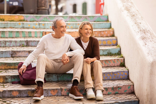 Boldog nyugdíjazást és vakációt. Vezető turisták pár ül a színes lépéseket a szabadban pihenés közben séta túra Európa városában. Érett utazók élvezik a városnézést - Fotó, kép