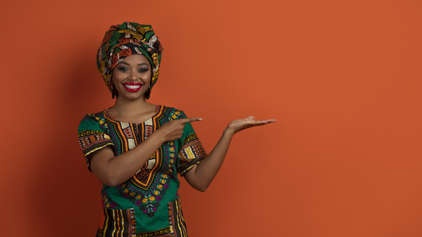 Ulusal kostümlü, mutlu, çekici bir Afrikalı kadın avucunda güzel bir şey gösteriyor ve gülümsüyor, fotokopi alanını işaret ediyor, turuncu stüdyo arka planında izole edilmiş, panorama. - Fotoğraf, Görsel