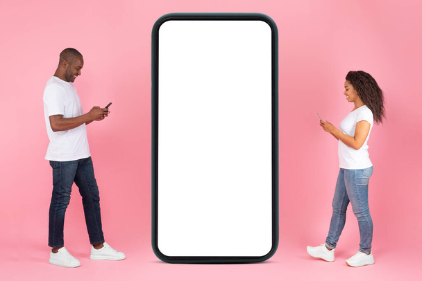 Modern online communication. Full length of positive black man and woman walking to big huge smartphone with white screen and using cellphones, chatting on social media mockup - Photo, Image