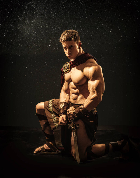 Bonito jovem muscular posando ajoelhado em trajes de gladiador romano ou espartano com escudo, em fundo preto no estúdio - Foto, Imagem