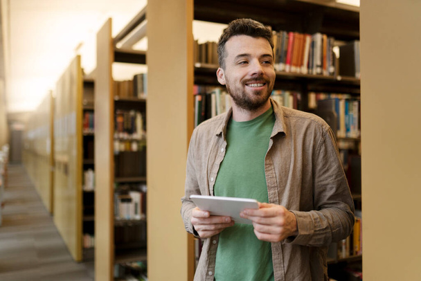 Glimlachende, knappe Spaanse man in de buurt van de boekenplank, met een digitale tablet, die e-boeken leest. Studenten van middelbare leeftijd die in de bibliotheek studeren, op zoek naar informatie - Foto, afbeelding