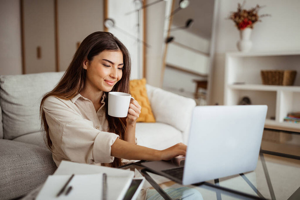 Joven mujer asiática alegre escribiendo en la computadora y beber taza de café en el interior de la habitación. Estudio, trabajo, negocios freelance en casa, nueva normalidad con tecnología, reuniones y redes sociales - Foto, Imagen
