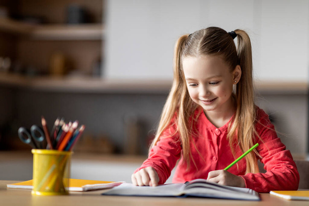 Hermosa niña preadolescente escribiendo con lápiz en un cuaderno mientras está sentada en el escritorio en casa, retrato de un lindo niño caucásico dibujando en un libro de trabajo, haciendo tarea escolar, espacio para copiar - Foto, Imagen