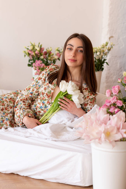 Mooie jonge vrouw in jurk met boeket van tulpen op het bed - Foto, afbeelding