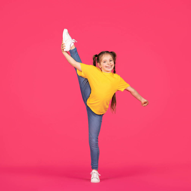 Linda niña gimnasta levantando su pierna en cuerda mientras posa sobre fondo de estudio rosa, niña preadolescente alegre en ropa casual haciendo gimnasia, haciendo ejercicios de estiramiento, longitud completa - Foto, Imagen