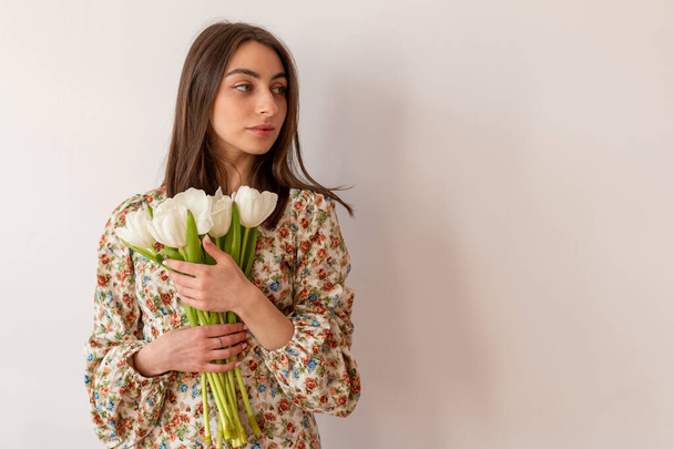 Pretty young woman in dress with flowers and dark hair with bouquet of tulips on the white wall background. - Фото, зображення