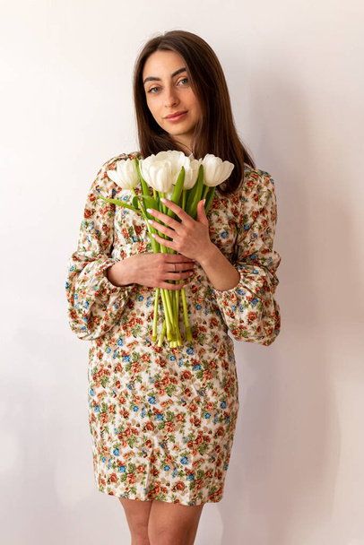 Mujer joven bonita en vestido con flores y cabello oscuro con ramo de tulipanes en el fondo blanco de la pared. - Foto, Imagen