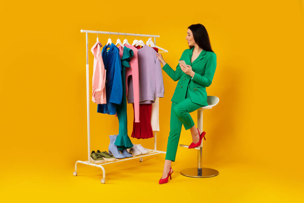 Jeune femme élégante tenant un téléphone portable et vérifiant de nouveaux vêtements, assise près du rail de vêtements sur fond de studio jaune, pleine longueur, espace de copie - Photo, image