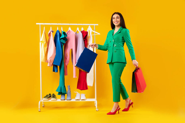 Shopping and seasonal sales. Happy stylish woman walking with shopper bags near clothing rail on yellow studio background, full length shot, free space - Photo, Image