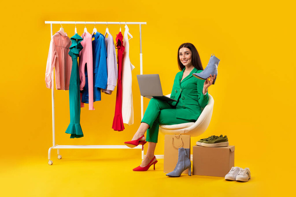 ラップトップを使用して興奮した女性スタイリストと新しいエレガントな靴を保持し、明るい服で衣料品レールの近くに座って、黄色のスタジオの背景にインターネット上で衣装を販売 - 写真・画像
