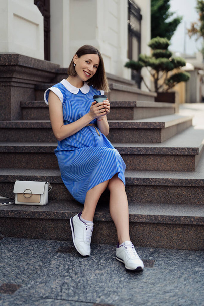 Pregnant woman in a blue dress touching her tummy while sitting and resting in the city on the stairs. Hands holding a cup with copy space - Fotó, kép