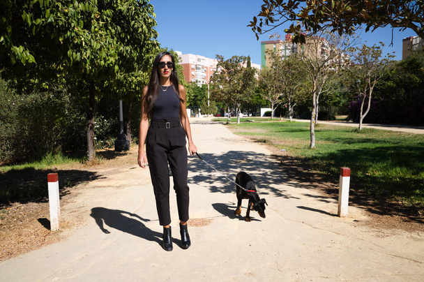 美しいとブルネットスペインの女性はドバーマンの子犬である彼女の犬を歩いています。女性はとてもエレガントな黒の服を着ている。ペットと動物のコンセプト. - 写真・画像