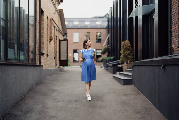 Απασχολημένη σύγχρονη έγκυος γυναίκα που περπατά ανάμεσα σε κτίρια γραφείων χρησιμοποιώντας ένα smartphone. Γυναίκα σε ένα μπλε vintage φόρεμα και λευκό πορτοφόλι χρησιμοποιώντας το τηλέφωνο στο δρόμο - Φωτογραφία, εικόνα