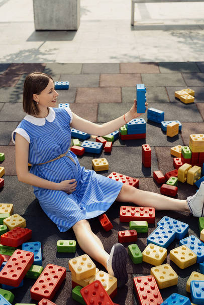 Es un niño. Una hermosa mujer embarazada en un vestido azul jugando con grandes cubos de colores en un parque infantil mientras se sienta y observa los cubos. Concepto creativo del embarazo - Foto, imagen