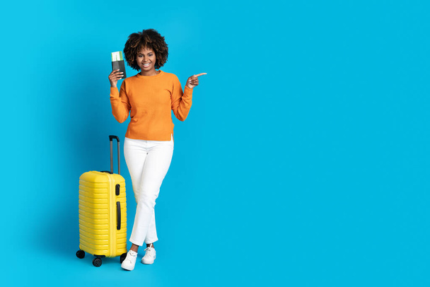 Cheery happy smiling pretty young black woman traveller carrying yellow luggage, holding passport and flight tickets, blue studio background, pointing at copy space, full length. Tourism, traveling - Фото, изображение