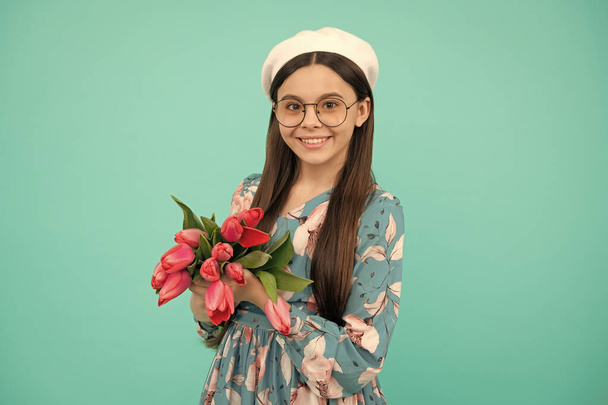 smiling child in beret with tulips. mothers or womens day. kid in glasses hold flowers for 8 of march. teen girl with spring bouquet on blue background. floral present. - Foto, Imagen