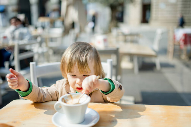 Cute toddler boy having hot chocolate in outdoor cafe. Small child drinking hot beverage on sunny terrace in Lecco. Spending good time on vacation with kids on Lake Como. Lecco, Lombardy, Italy. - Фото, зображення