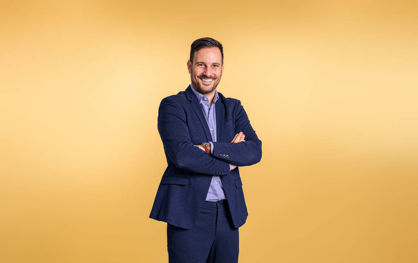 Confident handsome mid adult businessman wearing elegant blue suit with arms crossed smiling and posing on isolated orange background - Zdjęcie, obraz