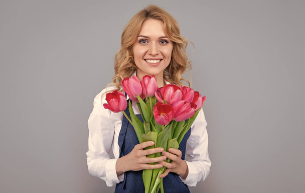 продавець квітів жінка посміхається в фартусі з весняними квітами тюльпанів на сірому фоні
. - Фото, зображення