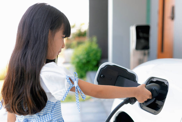 A playful girl holding an EV plug, a home charging station providing a sustainable power source for electric vehicles. Alternative energy for progressive lifestyle. - Fotó, kép