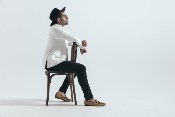 side view of cool fashion guy with hat and sunglasses sitting on wooden chair and looking to side in front of grey background in studio - Photo, Image