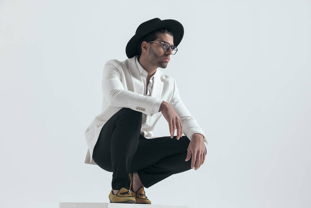 cool módní chlap s kloboukem a brýlemi drží paže na kolenou a krčí se před šedým pozadím ve studiu - Fotografie, Obrázek