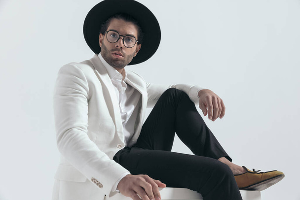 cool fashion man with hat and glasses sitting and holding elbow on knee in front of grey background - Photo, Image